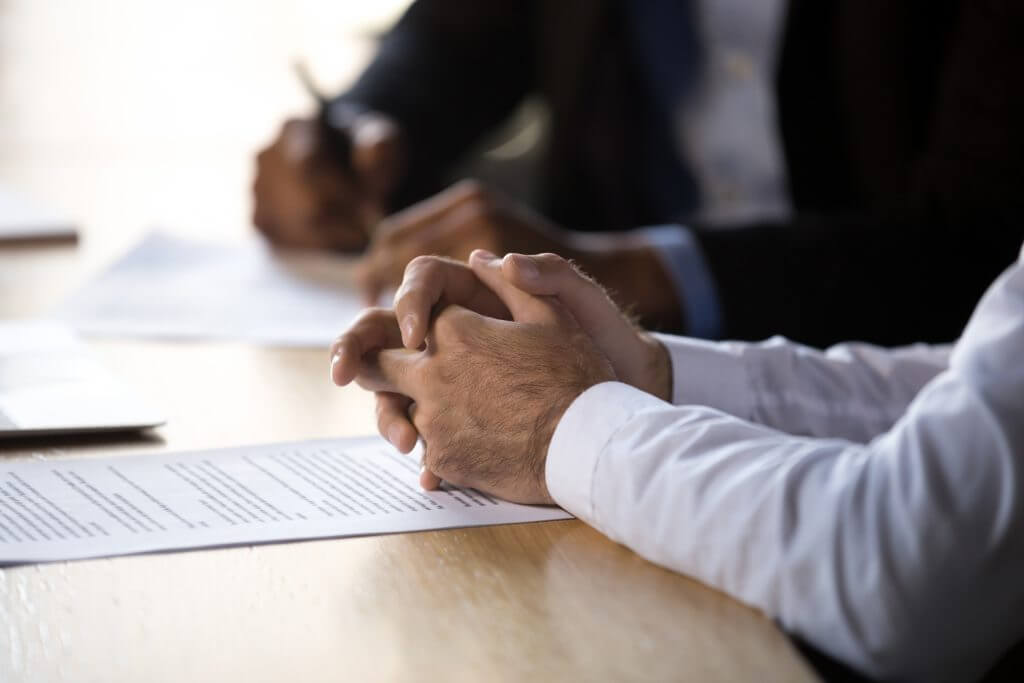Close-up of hands resting on a table with documents, two people in blurred background writing. Scene suggests a serious, professional meeting.